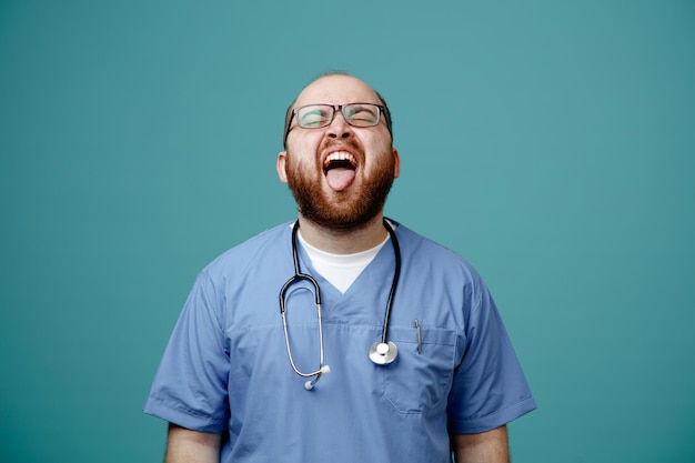 Бородатый мужчина-врач в форме со стетоскопом на шее в очках с высунутым языком и раздраженным выражением лица, стоящий на синем фоне