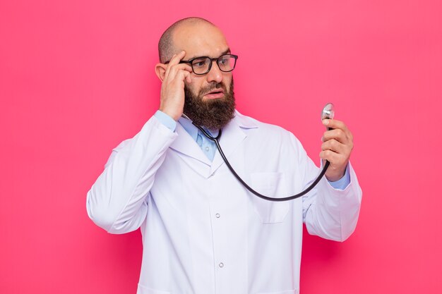 Бесплатное фото Бородатый мужчина-врач в белом халате в очках со стетоскопом смотрит на него изумленно, стоя на розовом фоне
