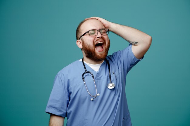 Bearded man doctor in uniform with stethoscope around neck wearing glasses happy and excited holding hand on his head standing over blue background