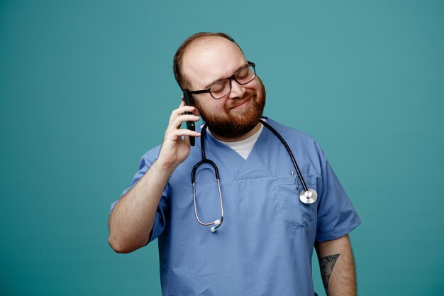 Бородатый мужчина-врач в форме со стетоскопом на шее в очках счастлив и доволен, разговаривая по мобильному телефону, улыбаясь, стоя на синем фоне