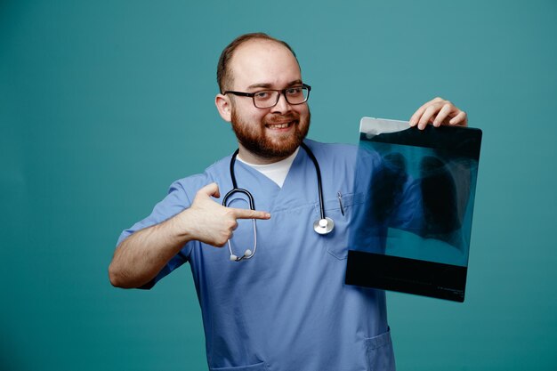 Bearded man doctor in uniform with stethoscope around neck wearing glasses holding lung xray happy and positive pointing with index finger at it smiling standing over blue background