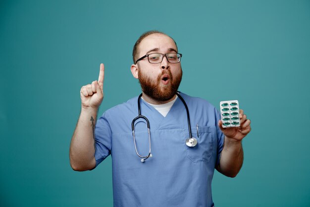 Bearded man doctor in uniform with stethoscope around neck wearing glasses holding pills looking at camera surprised showing index finger having great idea standing over blue background