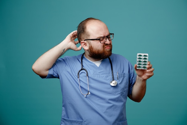 Бородатый мужчина-врач в униформе со стетоскопом на шее в очках с таблетками выглядит смущенным, почесывая голову, стоя на синем фоне