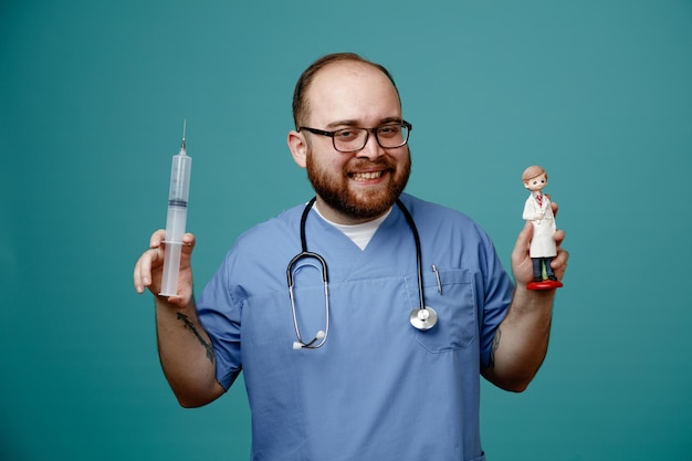 Бородатый мужчина-врач в форме со стетоскопом на шее в очках, держащий шприц и куклу, весело улыбающуюся, весело стоящую на синем фоне