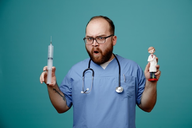 Bearded man doctor in uniform with stethoscope around neck wearing glasses holding syringe and a doll looking at camera frowning standing over blue background