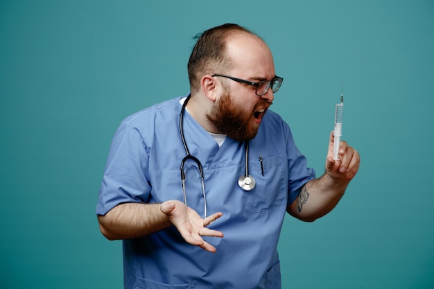 Bearded man doctor in uniform with stethoscope around neck wearing glasses holding syringe looking at it being displeased raising arm in displeasure standing over blue background