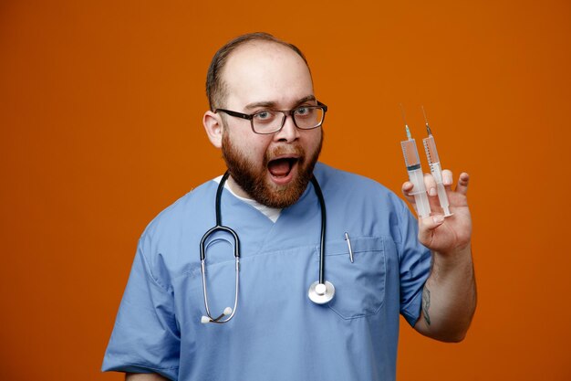 Bearded man doctor in uniform with stethoscope around neck wearing glasses holding syringes looking at camera happy and excited standing over orange background
