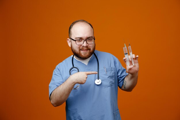 Bearded man doctor in uniform with stethoscope around neck wearing glasses holding syringes pointing with index finger at them smiling confident standing over orange background