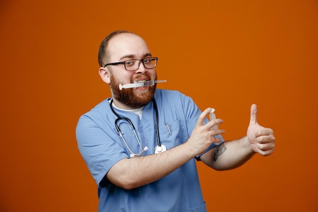 Uomo barbuto medico in uniforme con uno stetoscopio intorno al collo con gli occhiali che tengono le siringhe che mostrano il pollice in alto felice e gioioso in piedi su sfondo arancione