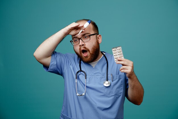 Bearded man doctor in uniform with stethoscope around neck wearing glasses holding thermometer and pills looking confused and worried standing over blue background