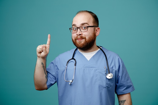Бородатый мужчина-врач в униформе со стетоскопом на шее в очках смотрит в камеру счастливым и позитивным, показывая указательным пальцем хорошие новости, стоя на синем фоне
