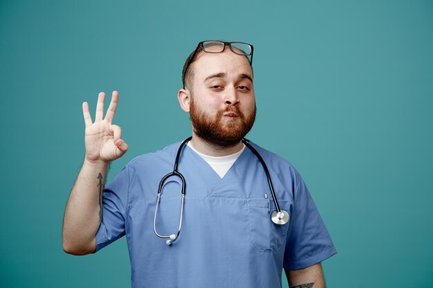 Bearded man doctor in uniform with stethoscope around neck wearing glasses looking at camera smiling confident showing ok sign standing over blue background