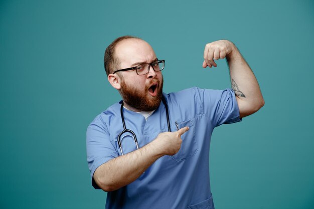 Bearded man doctor in uniform with stethoscope around neck wearing glasses looking confident and strong raising fist showing biceps standing over blue background