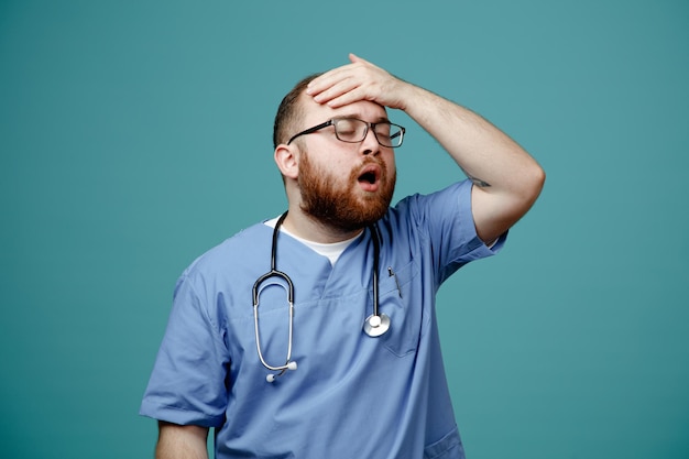 Бородатый мужчина-врач в форме со стетоскопом на шее в очках выглядит смущенным и разочарованным, держа руку на голове за ошибку, стоящую на синем фоне