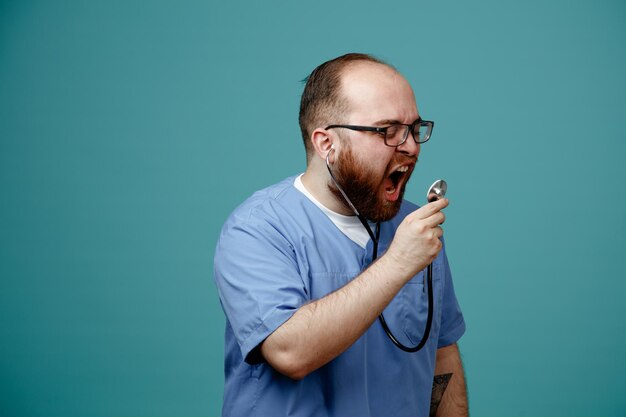 Бородатый мужчина-врач в форме со стетоскопом на шее в очках кричит в стетоскоп с агрессивным выражением лица, стоя на синем фоне