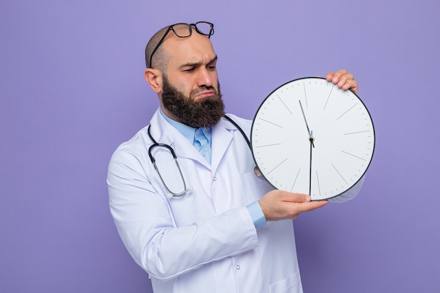 Бородатый мужчина-врач в белом халате со стетоскопом на шее держит часы, глядя на него недовольно и смущенно, стоя на фиолетовом фоне