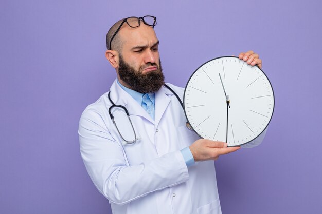 Bearded man doctor in white coat with stethoscope around neck holding clock looking at it displeased and confused standing over purple background