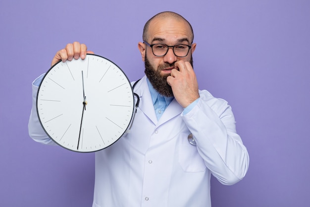 Bearded man doctor in white coat with stethoscope around neck holding clock looking with confuse expression