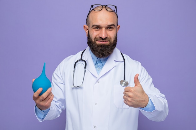 Uomo barbuto dottore in camice bianco con stetoscopio intorno al collo che tiene in mano una pera medica che sembra sorridente fiducioso che mostra i pollici in su