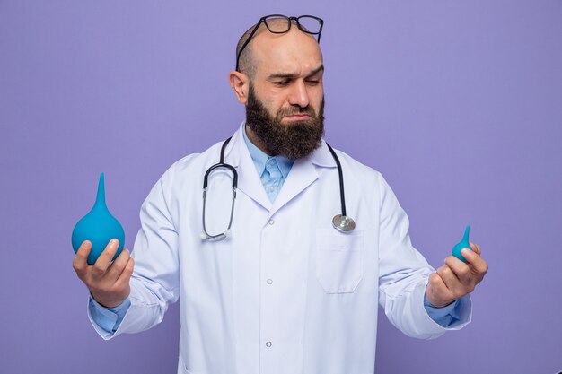 Bearded man doctor in white coat with stethoscope around neck holding medical pears looking confused trying to make choice standing over purple background