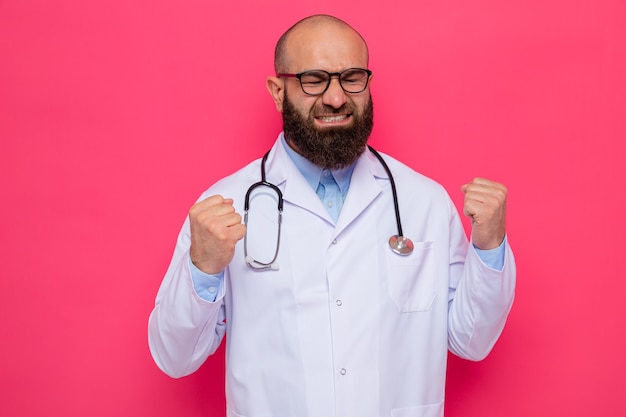 Bearded man doctor in white coat with stethoscope around neck wearing glasses clenching fists frustrated and angry standing over pink background