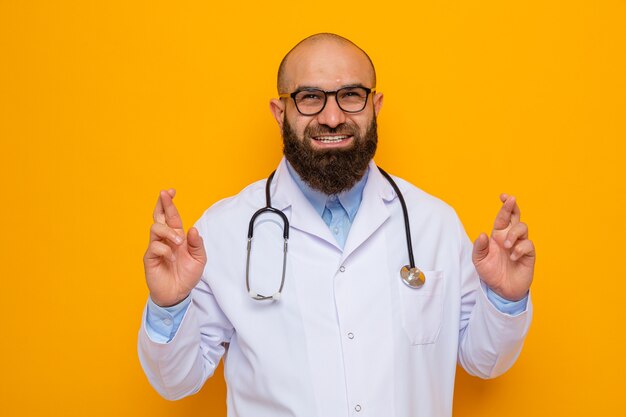 Bearded man doctor in white coat with stethoscope around neck wearing glasses, happy and cheerful making desirable wish crossing fingers