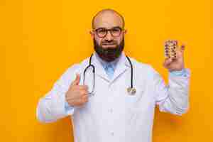 Foto gratuita uomo barbuto medico in camice bianco con stetoscopio intorno al collo con gli occhiali che tengono blister con pillole guardando la telecamera sorridendo allegramente mostrando i pollici in piedi su sfondo arancione