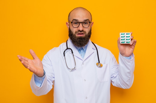 Bearded man doctor in white coat with stethoscope around neck wearing glasses holding blister with pills looking happy and surprised raising arm