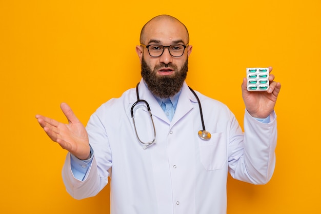 Бородатый мужчина-врач в белом халате со стетоскопом на шее в очках, держащий блистер с таблетками, выглядит счастливым и удивленным, поднимая руку
