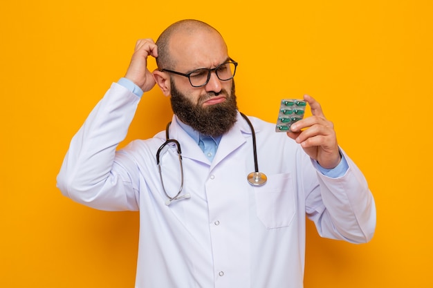 Bearded man doctor in white coat with stethoscope around neck wearing glasses holding blister with pills looking at it confused scratching head