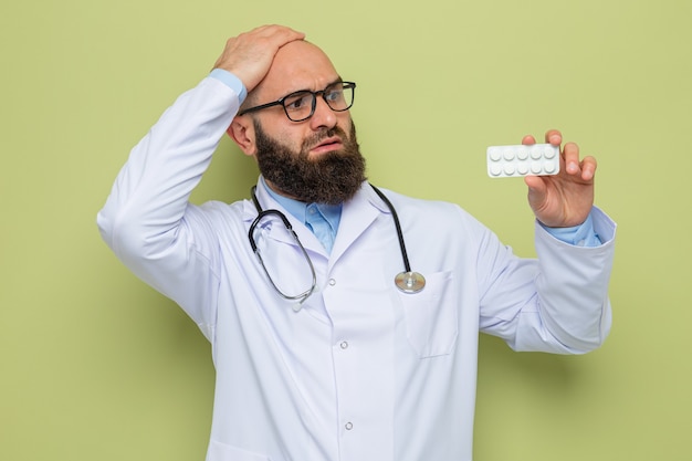 Medico uomo barbuto in camice bianco con stetoscopio intorno al collo con gli occhiali che tengono blister con pillole guardandolo confuso con la mano sulla testa