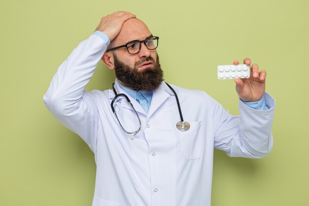 Бородатый мужчина-врач в белом халате со стетоскопом на шее в очках, держащий блистер с таблетками, глядя на него, смущенный рукой на голове