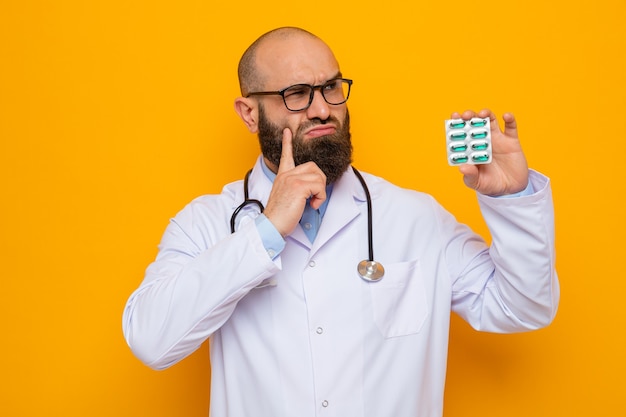 Bearded man doctor in white coat with stethoscope around neck wearing glasses holding blister with pills looking up puzzled