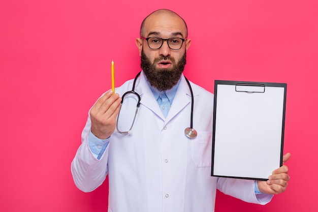 Bearded man doctor in white coat with stethoscope around neck wearing glasses holding clipboard with blank pages and pencil looking at camera surprised standing over pink background