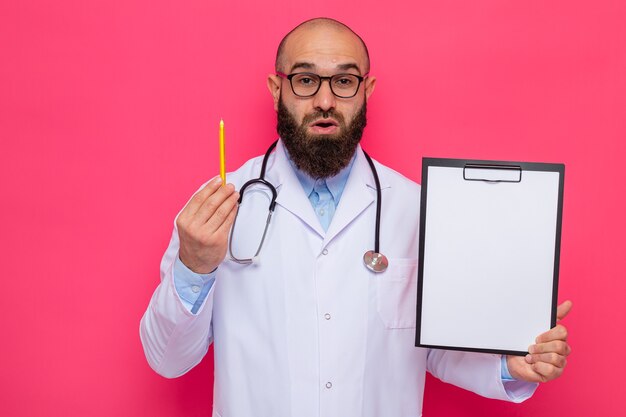 Bearded man doctor in white coat with stethoscope around neck wearing glasses holding clipboard with blank pages and pencil looking at camera surprised standing over pink background