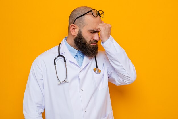 Bearded man doctor in white coat with stethoscope around neck wearing glasses holding fist on his forehead looking confused standing over orange background