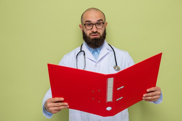 Bearded man doctor in white coat with stethoscope around neck wearing glasses holding office folder looking at camera with serious face standing over green background