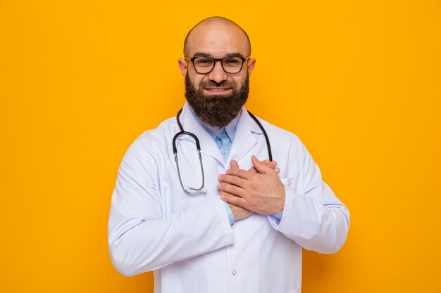 Bearded man doctor in white coat with stethoscope around neck wearing glasses looking at camera smiling cheerfully holding hands on his chest feeling thankful standing over orange background