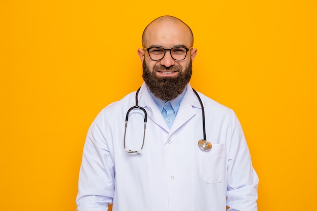 Bearded man doctor in white coat with stethoscope around neck wearing glasses looking at camera smiling confident standing over orange background