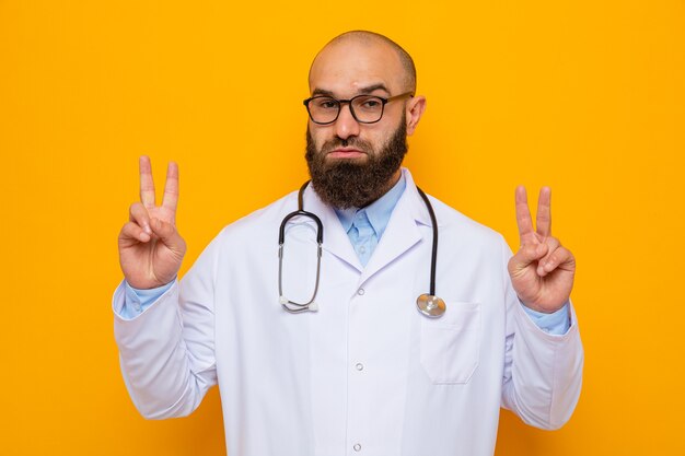 Bearded man doctor in white coat with stethoscope around neck wearing glasses looking at camera with confident expression showing v-sign standing over orange background