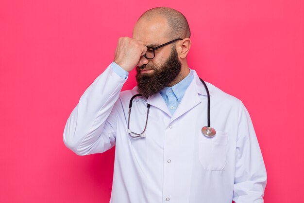 Free photo bearded man doctor in white coat with stethoscope around neck wearing glasses looking confused with fist on his forehead for mistake