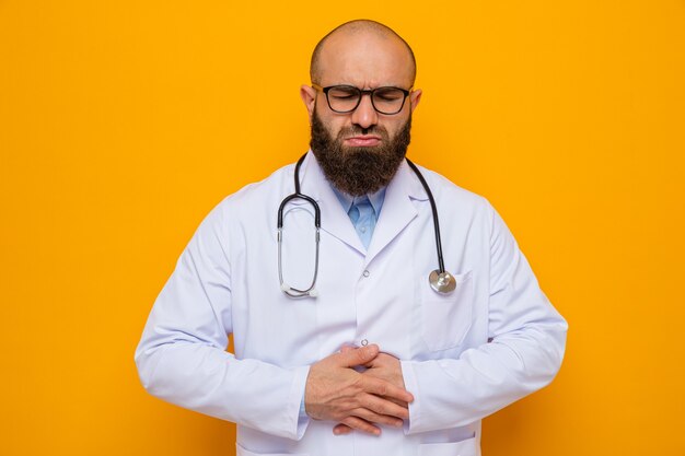 Bearded man doctor in white coat with stethoscope around neck wearing glasses looking unwell touching his belly feeling pain