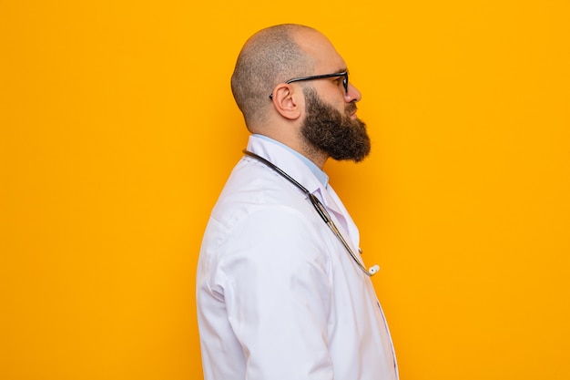 Uomo barbuto dottore in camice bianco con stetoscopio intorno al collo con gli occhiali in piedi di lato con faccia seria su sfondo arancione orange