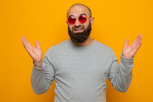 Bearded man in grey sweatshirt wearing red glasses looking happy and cheerful smiling broadly with arms raised
