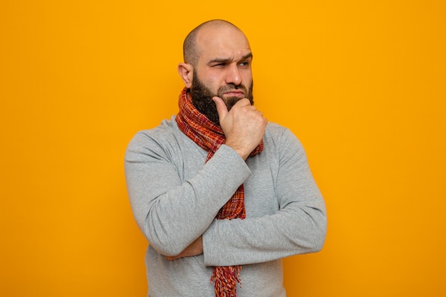 Bearded man in grey sweatshirt with scarf around his neck looking at side with pensive expression on face thinking