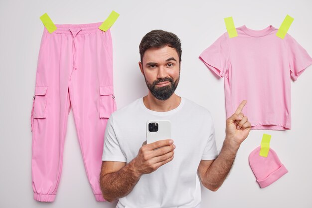 Free photo bearded man holds mobile phone checks newsfeed online indicates at hanging clothes suggests to buy outfit recommends something to wear isolated over white wall
