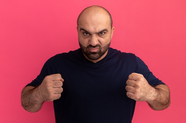 Бесплатное фото Бородатый мужчина в темно-синей футболке с серьезным сердитым лицом со сжатыми кулаками стоит над розовой стеной
