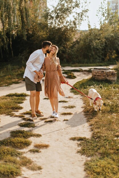 Bearded man kisses his girlfriend on cheek, holding her hand. Couple walking with labrador in garden.