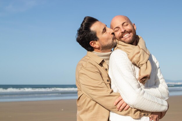 Bearded man kissing boyfriend on beach. Brunette middle-aged man with closed eyes hugging partner from back. Gays concept