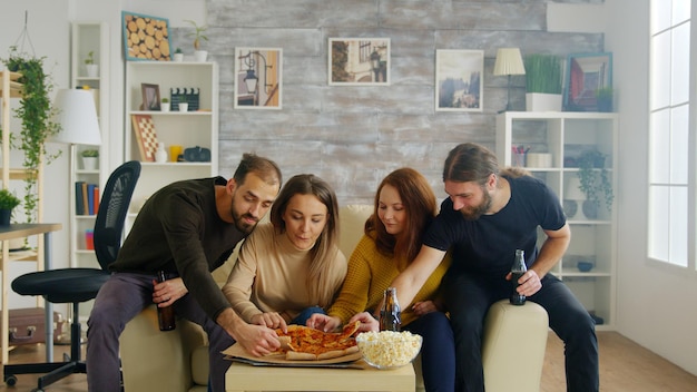 Bearded man laughing hard while watching tv with his friend and holding a beer bottle.